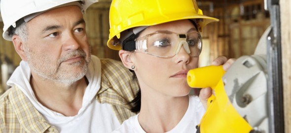 Female worker cutting wood with a power saw while male worker standing behind